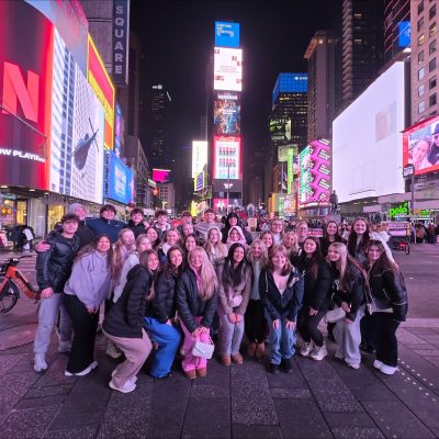 Group of teens posing for a photo in Times Square at night, with bright digital billboards and crowds in the background.