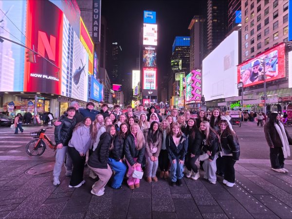 Group of teens posing for a photo in Times Square at night, with bright digital billboards and crowds in the background.