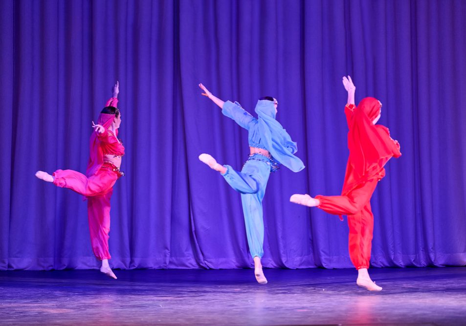 Vibrant red and blue costumes, dynamic odalisque designs, graceful leaps, theatrical stage with rich purple-blue curtain and soft warm spotlighting create exotic scheherazade atmosphere