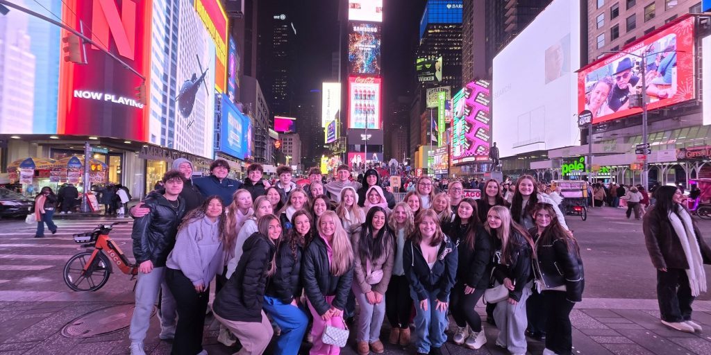 Group of teens posing for a photo in Times Square at night, with bright digital billboards and crowds in the background.